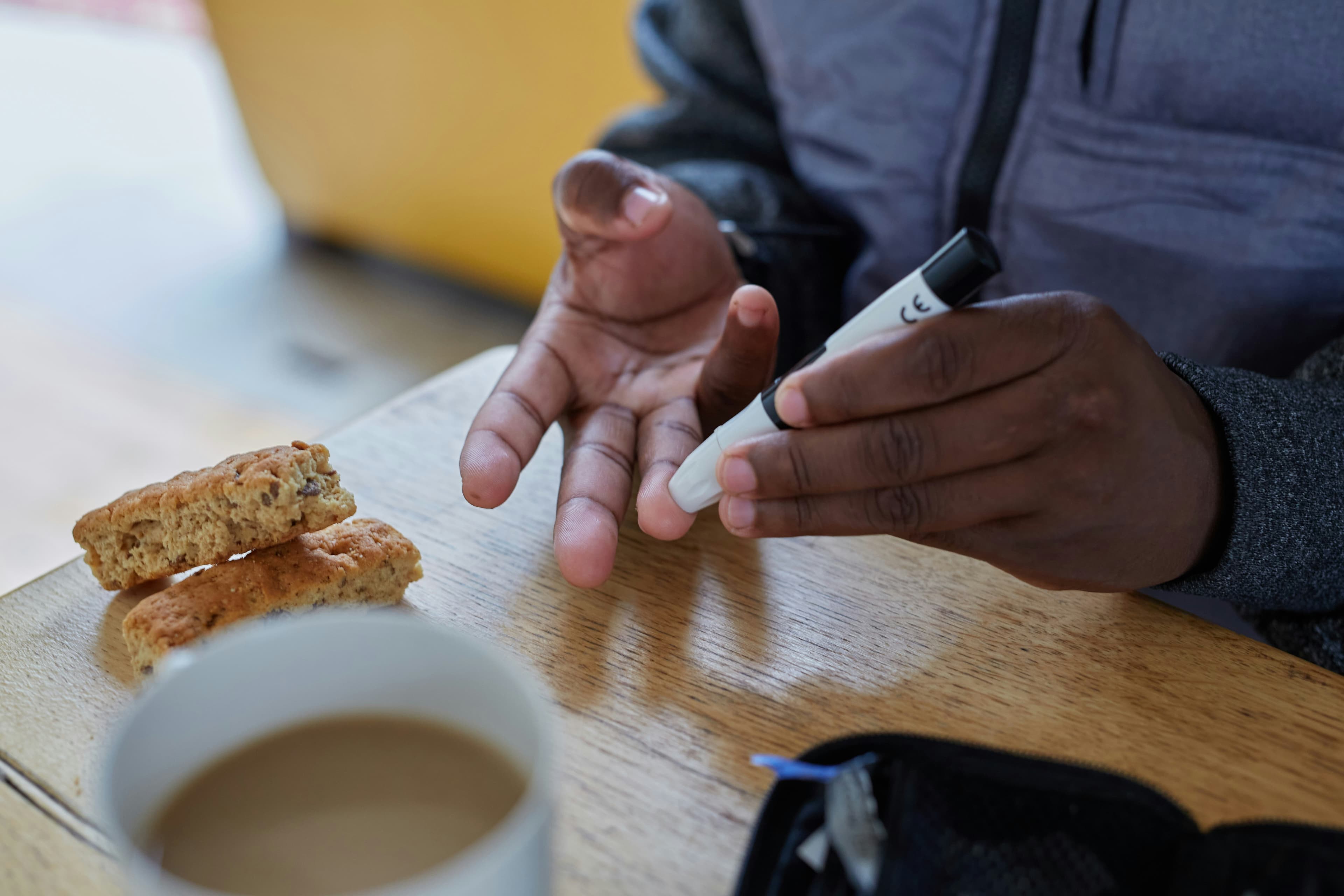 A diabetic checks their blood sugar with a lancing device at a café, coffee and a snack on the table.